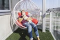 Millennial woman with white puppy sitting in modern hanging chair on balcony of apartment building and smile Royalty Free Stock Photo