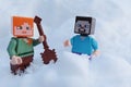 Minecraft LEGO figures of Steve and Alex standing in real deep snow, Alex starts digging snow with shovel.