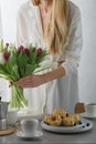 Mini bundt cakes with fruits on plate, cup of coffee and girl put bouquet of tulip flowers in a vase. Royalty Free Stock Photo