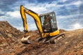 Mini excavator at the construction site on the edge of a pit against a cloudy blue sky. Compact construction equipment for Royalty Free Stock Photo