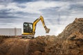 Mini excavator at the construction site on the edge of a pit against a cloudy blue sky. Compact construction equipment for Royalty Free Stock Photo