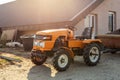 Mini red tractor with trailer standing near hangar building at farm during sunset or sunrise. Small agricultural