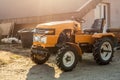 Mini red tractor with trailer standing near hangar building at farm during sunset or sunrise. Small agricultural