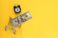 Mini shopping cart with 100 dollar banknotes inside and mini alarm clock on yellow background. Trolley and money. Financial crisis Royalty Free Stock Photo