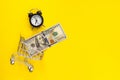 Mini shopping cart with 100 dollar banknotes inside and mini alarm clock on bright yellow background. Trolley and money Royalty Free Stock Photo
