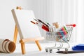 Mini shopping cart full with artistic goods and canvas on easel.