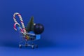 Mini shopping cart with small Christmas tree, Xmas decoration bauble balls and candy canes on navy blue background Royalty Free Stock Photo