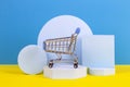 Mini shopping trolley cart with geometric podium platforms on yellow and light blue background Royalty Free Stock Photo