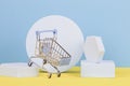 Mini shopping trolley cart with geometric podium platforms on yellow and light blue background. Online shopping concept Royalty Free Stock Photo