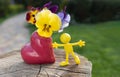 Miniature bright toy figurine of a yellow man near a heart-shaped vase with pansies