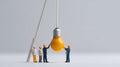 Miniature construction workers collaborating to install a large hanging yellow light bulb symbolizing teamwork and innovation in a Royalty Free Stock Photo