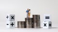 A miniature couple standing on a pile of coins with arithmetic symbols cube. Royalty Free Stock Photo