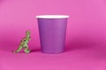 A miniature dinosaur and a paper cup against a purple background. A lilac disposable drink glass. Green figure of a predatory