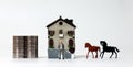 A miniature man standing in front of two piles of coins, a miniature house and two miniature horses.