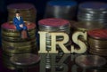 Miniature People. Man sitting on coin stacks with Text. IRS Taxman Concept. Macro