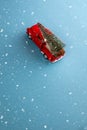 Miniature red car in the back carries a Christmas tree on a simple blue-aquamarine background under snow, top view. Save