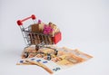 Miniature shopping cart filled with cash banknotes and a gifts boxes isolated on green background. Saving money for spending on