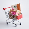 Miniature shopping cart filled with cash banknotes and a gifts boxes isolated on green background. Saving money for spending on
