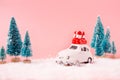 Miniature white car toy delivering gift box and christmas tree on pink background. Christmas greeting card concept Royalty Free Stock Photo