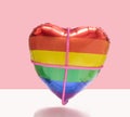 Minimal tight love concept. Rainbow color heart-shaped balloon with pink sparkle rope crosses it. Pink and star white background
