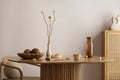 Minimalist composition of dining interior with round wooden table, design chair, vase with dried flowers, coconut in bowl,