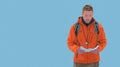 Adventurous Man in Orange Jacket Reads Map with Backpack and Compass, Blue Background Royalty Free Stock Photo