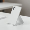 A minimalist, white smartphone is positioned on a sleek, triangular stand on a desk. The