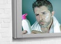 Mirror reflection portrait of young confused attractive man using beauty facial beauty product applying cream and reading lotion b Royalty Free Stock Photo