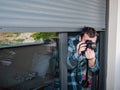 Man in a robe takes photos with a reflex camera from the balcony under the blind