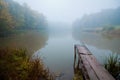 Misty autumn morning on a small lake, fog weather, wooden footbridge over still water surface, yellow trees reflections Royalty Free Stock Photo