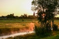 Misty stream winds through a glowing meadow at sunrise sunset, with a church spire silhouetted against the bright, warm sky. Royalty Free Stock Photo
