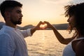 Mixed race couple, Caucasian man with her Hispanic girlfriend, forming a heart with their hands framing the setting sun on a lake