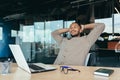 mixed race man relaxing in office with hands behind head looking out window and smiling, businessman daydreaming inside