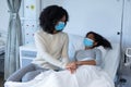 Mixed race mother comforting ill daughter in hospital bed, holding hands, both wearing face masks Royalty Free Stock Photo