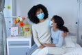 Mixed race mother embracing ill daughter sitting on hospital bed, both wearing face masks Royalty Free Stock Photo