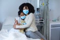 Mixed race mother hugging sick daughter in hospital bed holding teddy bear, all wearing face masks Royalty Free Stock Photo