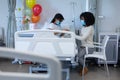 Mixed race mother and sick daughter in face masks in hospital, balloons next to them