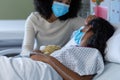 Mixed race mother and sick daughter in face masks in hospital, girl sleeping and holding teddy bear Royalty Free Stock Photo