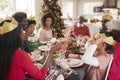 Mixed race, multi generation family sitting at the Christmas dinner table reading jokes and wearing paper party hats from Christma