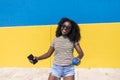 Mixed race smiling black woman portrait with big afro curly hair against blue and yellow wall dancing while holding a smartphone Royalty Free Stock Photo