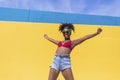 Mixed race smiling black woman portrait with big afro curly hair against blue and yellow wall dancing while holding a smartphone Royalty Free Stock Photo