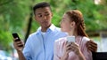 Mixed-race teen couple looking at each other chatting on smartphone in park Royalty Free Stock Photo