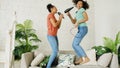 Mixed race young funny girls dance singing with hairdryer and comb jumping on sofa. Sisters having fun leisure in living