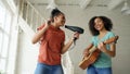 Mixed race young funny girls dance singing with hairdryer and playing acoustic guitar on a bed. Sisters having fun Royalty Free Stock Photo