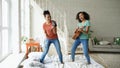 Mixed race young funny girls dance singing with hairdryer and playing acoustic guitar on a bed. Sisters having fun Royalty Free Stock Photo