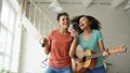 Mixed race young funny girls dance singing with hairdryer and playing acoustic guitar on a bed. Sisters having fun