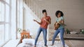 Mixed race young funny girls dancing singing and playing acoustic guitar on a bed. Sisters having fun leisure in bedroom