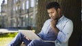 Mixed-race young man sitting under tree, looking at laptop, important letter Royalty Free Stock Photo