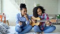 Mixed race young woman with tablet computer sitting on bed teaching her teenage sister to play acoustic guitar at home Royalty Free Stock Photo