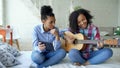 Mixed race young woman with tablet computer sitting on bed teaching her teenage sister to play acoustic guitar at home Royalty Free Stock Photo